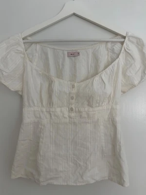 Vit blus med puffärm från Nelly - Söt vit blus från Nelly med puffärmar och knappar framtill. Blusen har en lätt rynkad struktur, markerad midja och rundad halsringning. Perfekt till jeans eller kjol för en fräsch look.