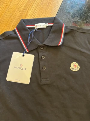 Svart Moncler pikétröja  - Klassisk svart pikétröja från Moncler med röd och vit rand på krage och ärmslut. Tröjan har knappslå med tre knappar och Moncler-logga broderad på bröstet. Tillverkad i bomull för en bekväm känsla.