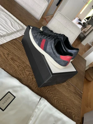 Gucci ace svarta - Snygga svarta Gucci sneakers med klassiskt GG-mönster och röda samt blå ränder på sidan. Skorna har vita sulor och är låga i modellen. Materialet är en mix av canvas och läder, vilket ger en exklusiv känsla. Perfekta för dig som vill sticka ut med stil. Storlek 42 du kan skriva för mer bilder eller om du har frågor😁