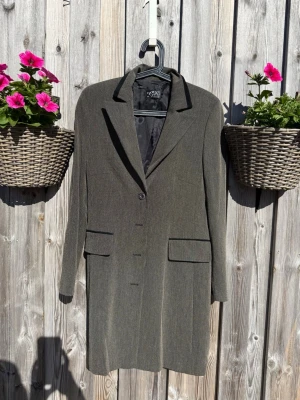 Klassisk gråbrun kappa från GM Design (S) - Stilren grå kappa från GM Design i storlek 36. Kappan har en rak passform, klassisk krage och knäppning framtill med fyra knappar. Två fickor med lock ger en snygg detalj. Materialet är tunt och har en diskret struktur som ger ett elegant intryck.