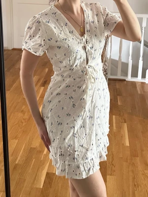 Vit blommig kortklänning med snörning - Supersöt vit kortklänning med små blå blommor, v-ringning och snörning framtill. Klänningen har volangkant nertill och korta ärmar. Perfekt för sommarens alla tillfällen. Passar mer som S.