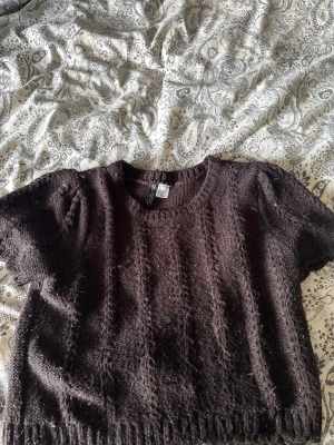 Mörkbrun stickad topp H&M - Kortärmad stickad topp från H&M i mörkbrunt garn. Tröjan har rund halsringning, puffiga ärmar och ribbstickad kant nedtill. Materialet ser mjukt och lite luddigt ut, perfekt för lager på lager och en trendig look.