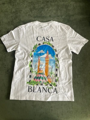 Vit Casablanca t-shirt med tryck - Vit t-shirt från Casablanca med färgglatt tryck av byggnader och palmer på ryggen samt mindre tryck på bröstet. Klassisk rund hals och korta ärmar. Perfekt för dig som gillar stilrena plagg med unik design.