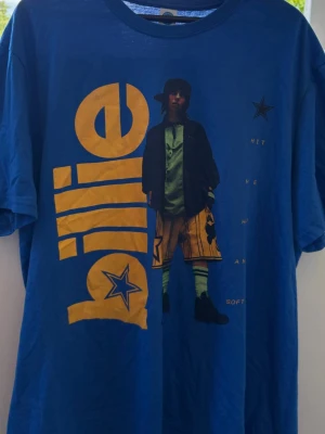 Blå billie Eilish t-shirt - Blå Billie Eilish t-shirt med stort gult tryck av texten 'billie' och en grafisk bild av Billie Eilish på framsidan. T-shirten har korta ärmar och en avslappnad passform. Den är aldrig använd! 