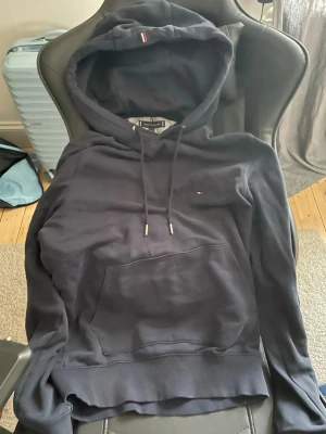 Mörkblå hoodie från Tommy Hilfiger - Snygg mörkblå hoodie från Tommy Hilfiger med klassisk huva och dragsnören. Liten logga på bröstet och en diskret detalj på huvan. Stor magficka framtill och ribbade muddar vid ärmslut och nederkant. Perfekt för en avslappnad och stilren look.