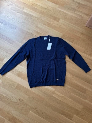 Mörkblå strukturerad tröja från SAMS - Mörkblå tröja från SAMS i storlek XL med snyggt strukturerat mönster. Tröjan har rund halsringning och ribbade muddar vid ärmslut och nederkant. Perfekt för dig som gillar stilrena och moderna plagg.