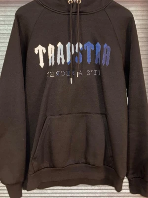 Svart Trapstar hoodie med tryck - Säljer en svart hoodie från Trapstar med stor ficka framtill och huva med dragsko. Framsidan har Trapstar-loggan i vitt och blått samt texten 'It's a secret'. Tillverkad i mjukt bomullsmaterial, perfekt för en chill och streetwear-inspirerad look.