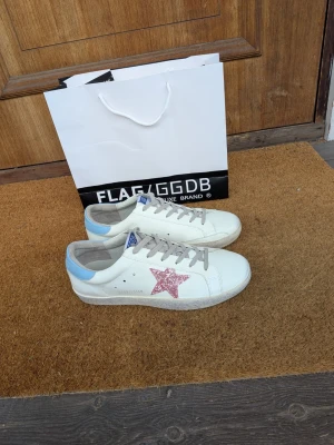Golden Goose vita sneakers med rosa stjärna - Golden Goose sneakers i vitt läder med en glittrig rosa stjärna på sidan och ljusblå detalj vid hälen. Skorna har grå skosnören och blå GGDB-märke på plösen. Klassisk låg modell med slitstark sula och trendig design.