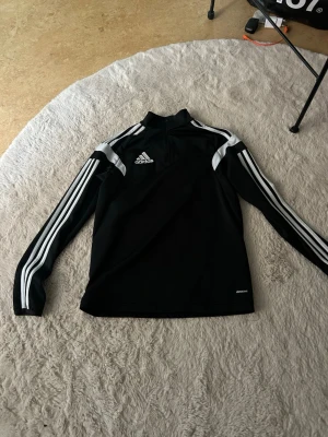 Svart Adidas träningströja YXL - Svart långärmad träningströja från Adidas med vita ränder längs ärmarna och vit logga på bröstet. Tröjan har halv dragkedja vid halsen och är tillverkad i funktionsmaterial. Perfekt för träning eller sportaktiviteter. Passar XXS/XS