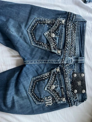 Blå Miss Me jeans med strassdetaljer - Snygga blå bootcut jeans från Miss Me med coola strassdetaljer och dekorativa sömmar på både fram- och bakfickor. Jeansen har låg midja, klassisk femficksmodell och silverfärgade knappar med logga. Perfekta för dig som gillar att sticka ut med din stil. När jag köpte dem så var de uppsydda med en kil, jag har sprätt upp dem längst ner för att göra de längre. Små i storleken. 💕