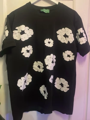 Svart t-shirt med vita blommor - Cool svart t-shirt med stora vita blommor tryckta över hela plagget. T-shirten har klassisk passform och korta ärmar, perfekt för dig som vill sticka ut med ett unikt mönster. Materialet känns mjukt och bekvämt mot huden.