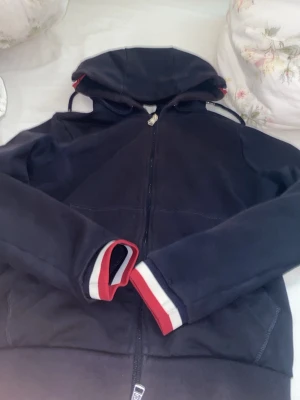 Moncler zip - Snygg mörkblå hoodie från Moncler med dragkedja, huva och dragsko. Röd, vit och blå randdetalj på huva och ärmslut. Klassisk Moncler-logga på ena ärmen. Perfekt för en chill och sportig stil. lite solbränd vid luvan
