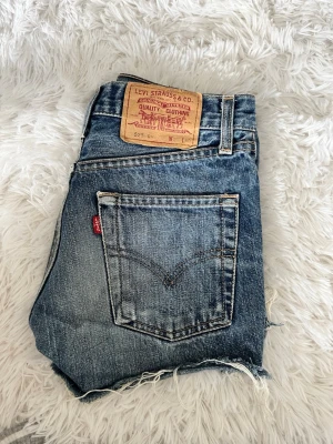 Levi's blå jeansshorts med rå kant - Säljer ett par klassiska blå jeansshorts från Levi's med rå, fransig kant och bakficka med den ikoniska röda Levi's-lappen. Shortsen har normal passform och är perfekta för sommaren. Snygg vintagekänsla och tidlös design.