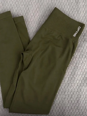 Olivgröna leggings från DFyne - Snygga olivgröna leggings från Dfyne med hög midja och bred ribbad linning. Tillverkade i ett stretchigt material som sitter skönt och formar sig efter kroppen. Perfekta för träning eller chill dagar. Diskret logga på linningen.