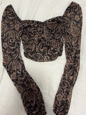 Svart blus med paisleymönster - Trendig svart blus med beige och brunt paisleymönster. Modellen är croppad med långa puffärmar och lätt v-ringad hals. Tunn, luftig polyester som ger en snygg look. Perfekt för dig som vill sticka ut med coola detaljer och unik design.