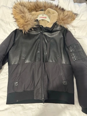 Svart pufferjacka med fuskpäls - Säljer en svart pufferjacka från Steam Park med detaljer i fejkläder och en stor huva med beige fuskpäls. Jackan har dragkedja framtill, fickor med dragkedja och ribbade muddar. Insidan av huvan är fodrad med mjuk teddy. Perfekt för kalla vinterdagar.