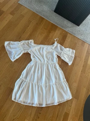 Vit offshoulder volangklänning - Superfin vit klänning med offshoulder och vida, volangiga ärmar. Klänningen har flera volanglager och resår i midjan för en snygg passform. Perfekt för sommarens alla tillfällen. Materialet känns lätt och luftigt.