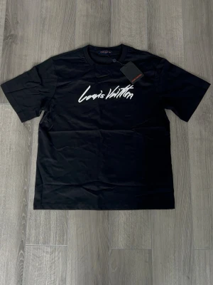 Svart Louis Vuitton t-shirt - Snygg svart t-shirt från Louis Vuitton med vit logga i grafisk stil på bröstet. Klassisk rund halsringning och korta ärmar. Tillverkad i mjuk bomull som känns skön mot huden. Perfekt för dig som gillar exklusiva streetwear-vibbar.