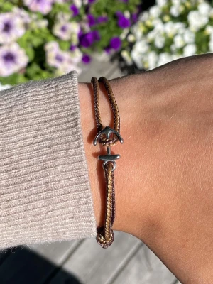 Brunt armband med ankare - Snyggt armband i brunt snöre med ett silverfärgat ankare som låsdetalj. Armbandet har en enkel och stilren design som passar perfekt till vardagslooken. Justerbar storlek tack vare knutarna på sidorna.