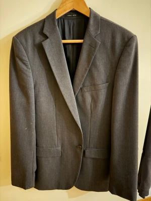 Grå kavaj från Zara Man - Stilren grå kavaj från Zara Man med klassisk passform och enkelknäppning. Kavajen har två framfickor med lock, bröstficka och smala slag. Perfekt för en dressad look. Materialet är slätt och ger ett elegant intryck. Storlek 48