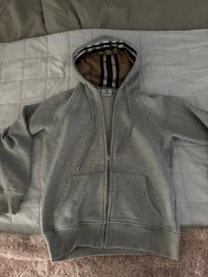 Grå zip-hoodie med mönstrad huva - Snygg grå hoodie med dragkedja. Klassisk huva med det ikoniska rutmönstret på insidan, dragsko och två stora fickor framtill. Perfekt för en chill och stilren look. Mjuk bomullskänsla och ribbade muddar vid ärmslut och nederkant.