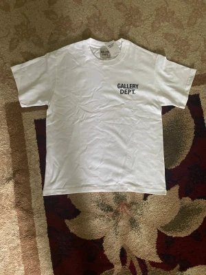  Gallery Dept - Vit t-shirt från Gallery Dept. med svart tryck på bröstet och stort tryck på ryggen med texten 'Gallery Dept. Hollywood, CA.'. Klassisk passform och rund halsringning. Perfekt för dig som gillar streetwear och enkel stil.