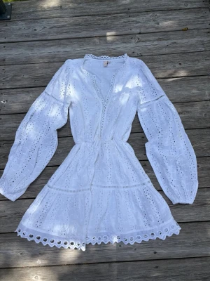 Vit kort klänning med broderi - Superfin vit kort klänning med broderade detaljer och volangkant. Klänningen har långärmade ärmar, v-ringning och markerad midja med knytband. Perfekt för sommarens alla tillfällen och ger en romantisk vibe.
