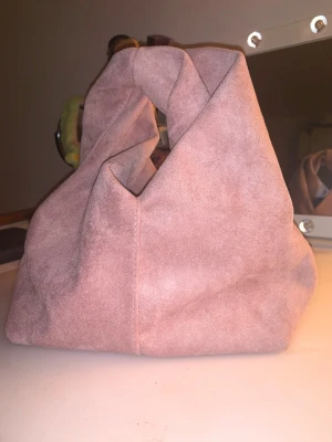 Rosa mocka handväska med unik form - Säljer en snygg handväska i mjuk rosa mocka med en cool, asymmetrisk design. Väskan har ett stort huvudfack och stängs med magnetknapp. Perfekt för dig som gillar trendiga accessoarer och vill sticka ut lite extra. Har aldrig använts! 