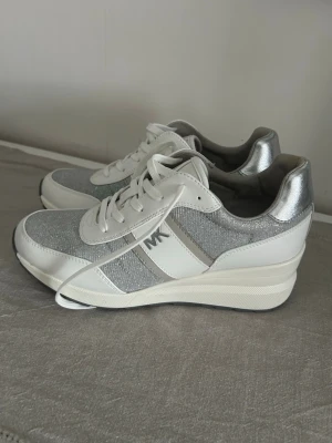 Vita och silver sneakers från Michael Kors - Snygga sneakers från Michael Kors i vitt och silver med glittrande textil och detaljer i skinnimitation. Klassisk låg modell med snörning och diskret MK-logga på sidan. Perfekta för dig som vill ha en stilren och trendig look.