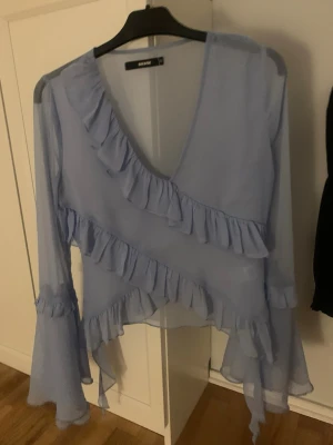 Ljusblå volangblus från Bik Bok - Supersöt ljusblå blus från Bik Bok med djup V-ringning och volangdetaljer framtill och på ärmarna. Blusen är gjord i ett tunt, transparent tyg och har långa, utsvängda ärmar som ger en romantisk känsla. Perfekt att styla med jeans eller kjol.
