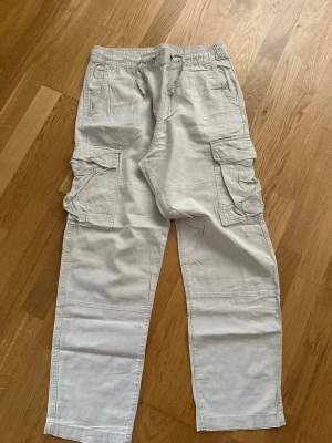 Beiga linnebyxor från H&M - Säljer ett par beiga linnebyxor från H&M med regular fit. Byxorna har dragsko i midjan, stora cargofickor på sidorna och är tillverkade i en mix av lin och bomull. Perfekta för en avslappnad och trendig look.
