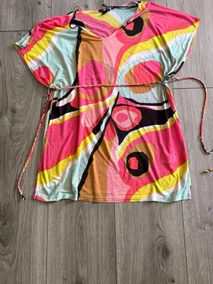 Färgglad kortklänning från H&M - Säljer en färgglad kortklänning från H&M med abstrakt mönster i rosa, gul, svart, beige och ljusblå. Klänningen har v-ringning, korta ärmar och ett knytband i midjan för en snygg passform. Perfekt för sommarens alla tillfällen!