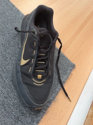 Svarta Nike Air Max sneakers - Snygga svarta Nike Air Max sneakers med guldfärgade detaljer och mesh-överdel för bra ventilation. Skon har en synlig Air Max-sula och klassisk Nike Swoosh på sidan. Perfekt för dig som gillar sportig och stilren look.