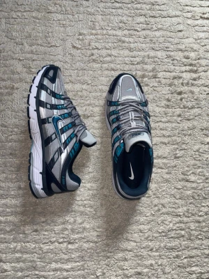 Nike p6000 - Snygga Nike sneakers i silver, blått och svart med mesh- och syntetmaterial. Skorna har en sportig design med tydliga detaljer och en robust sula med blå inslag. Perfekta för dig som gillar streetwear och vill sticka ut med coola färgkombos.