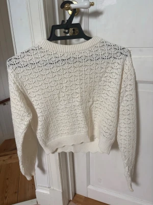 Vit virkad stickad tröja - Säljer en vit stickad tröja med virkat mönster och rund hals. Tröjan har långa ärmar och ribbade muddar vid ärmslut och nederkant. Perfekt för lager på lager och ger en söt, luftig look.