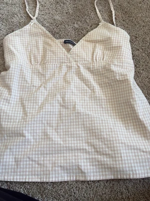 Beige rutig linnetopp från Brandy Melville - Söt beige och vit rutig linnetopp från Brandy Melville med smala axelband och v-ringning. Toppen har knyt i ryggen för extra detalj och sitter snyggt på kroppen. Perfekt till sommaren och enkel att matcha med jeans eller kjol.