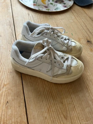 Vita New Balance sneakers med beige detaljer - Snygga vita New Balance sneakers med beige detaljer och tjock sula. Skorna har klassisk låg modell, snörning och blandning av läder och mocka i material. Perfekt för dig som gillar stilrena och sportiga sneakers.