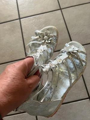 Vita sandaler med blommor storlek 36 - Säljer ett par vita sandaler med flera smala remmar och dekorativa blommor framtill. Sandalerna har en lätt genomskinlig sula med textmönster och rosa detaljer undertill. Perfekta för sommarens alla äventyr!