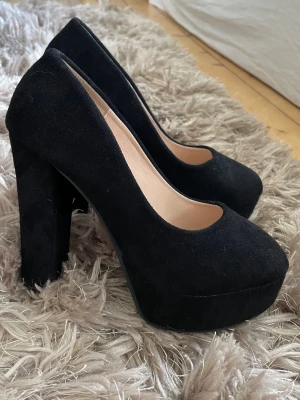 Svarta platåpumps med chunky klack - Säljer ett par svarta pumps med hög platå och chunky klack. Skorna har rundad tå och är klädda i mockaimitation för en snygg och trendig look. Perfekta för dig som vill ha extra höjd och stabilitet. Innersulan är beige och skon har en klassisk siluett.