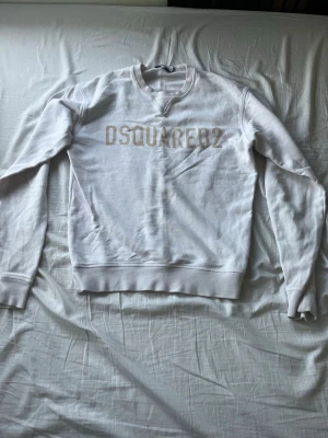 Vit sweatshirt från Dsquared2 - Säljer en vit sweatshirt från Dsquared2 med klassisk logga i beige på bröstet. Tröjan har rund halsringning, långa ärmar och ribbade muddar vid ärmslut och nederkant. Perfekt för dig som gillar stilrena och trendiga plagg. Priset kan såklart alltid diskuteras