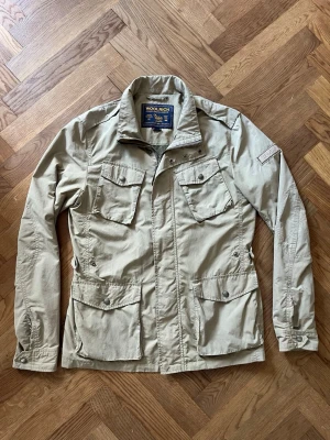 Beige parkajacka från Woolrich - Stilren beige vindjacka från Woolrich med fyra stora fickor framtill och hög krage. Jackan är tillverkad i bomull och polyester, har långa ärmar och stängs med dragkedja och knappar. Perfekt för dig som gillar klassisk och funktionell stil.