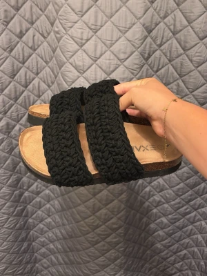 Svarta sandaler med stickade band - Säljer ett par svarta sandaler med två breda stickade band över foten och en bekväm, ljus innersula. Sulan är något upphöjd och har en klassisk slip-in design. Perfekta för en avslappnad och trendig look.