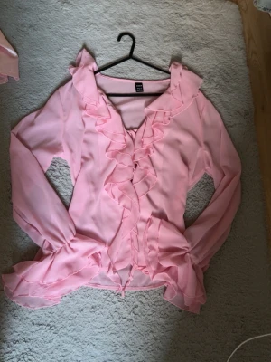 Rosa volangblus med knytband - Supersöt ljusrosa blus med volangdetaljer längs halsringningen och framtill. Blusen har långa ärmar och knytband under. Tillverkad i ett tunt, lätt och transparent tyg som ger en luftig känsla.