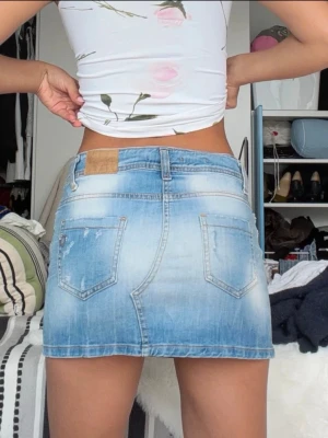 Blå lowrise jeanskjol i kort modell från Please - Snygg ljusblå jeanskjol i kort modell med klassiska fickor fram och bak samt bälteshällor. Kjolen har en lätt tvättad look och stängs med knapp och dragkedja framtill. Perfekt för en avslappnad och trendig stil.