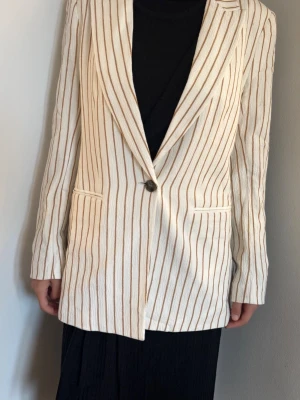 Randig beige kavaj med bruna ränder - Snygg beige kavaj med tunna bruna vertikala ränder och en knapp framtill. Kavajen har två fickor, klassisk krage och är tillverkad i ett strukturerat tyg. Perfekt för dig som vill ha en trendig och stilren look. OBS! Modellen är XS och bär storlek S (38)