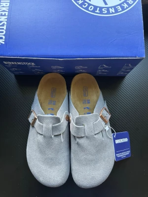 Grå mockatofflor från Birkenstock - Snygga grå tofflor från Birkenstock i mjuk mocka med justerbart spänne i metall. Klassisk slip-in modell med bekväm fotbädd och korksula. Perfekta för dig som vill ha både stil och komfort i vardagen.