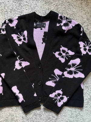 Svart kofta med lila fjärilar ASOS - Snygg svart kofta från ASOS DESIGN med lila fjärilsmönster. Koftan har v-ringning, tre knappar framtill och ribbade muddar. Perfekt för dig som gillar statement-plagg och vill ha något unikt i garderoben.