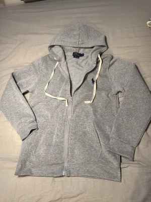Grå hoodie från Polo Ralph Lauren - Snygg grå hoodie från Polo Ralph Lauren med dragkedja, huva och vita snören. Klassisk broderad logga i mörkblått på bröstet. Mjuk bomullsblandning och två fickor framtill. Perfekt för en chill och stilren look.
