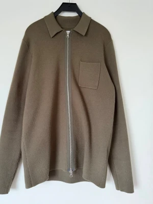 Olivgrön overshirt från Samsøe Samsøe - Stilren olivgrön overshirt från Samsøe Samsøe med rak passform, klassisk krage och silverfärgad dragkedja framtill. En bröstficka på vänster sida och mjukt, tjockt material som känns lyxigt. Perfekt lager på lager-plagg för en trendig look.