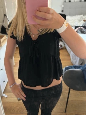 Svart blus med volang och knyt - Säljer en svart blus med volangdetaljer vid ärmarna och knyt framtill. Blusen har v-ringning, korta ärmar och är lätt croppad framtill. Perfekt till jeans eller kjol för en trendig look.